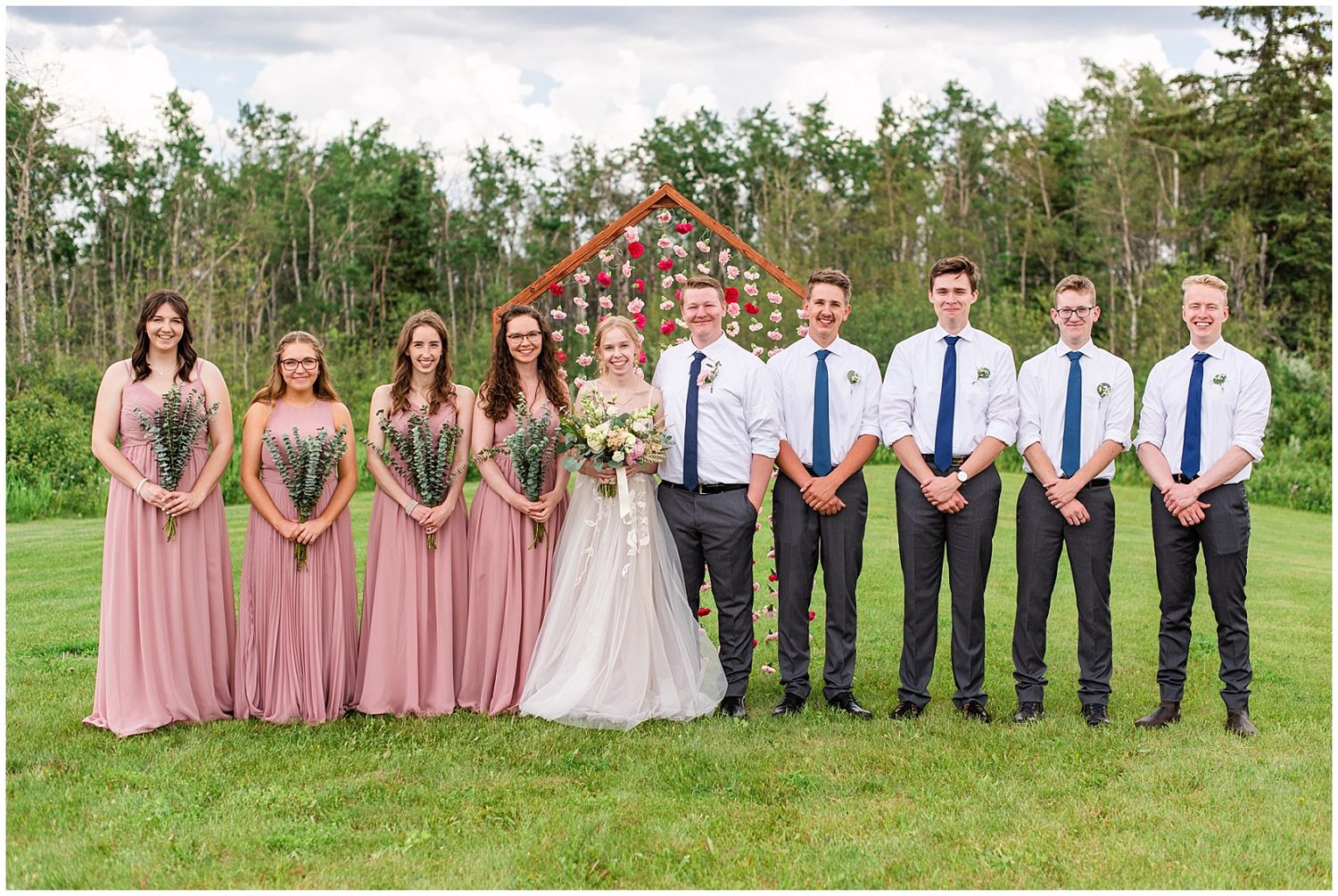 Wedding party poses outdoors with bridesmaids in dusty rose gowns and groomsmen in white shirts with navy ties before a floral arch.
