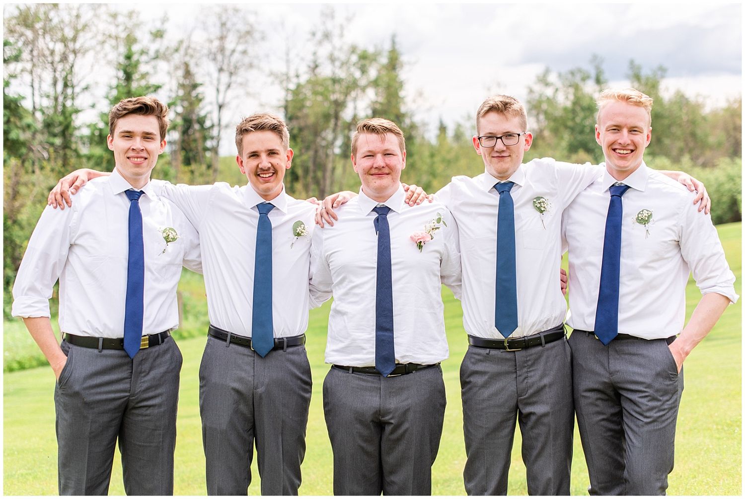 Five groomsmen in white shirts and blue ties pose together smiling on a green lawn outdoors.