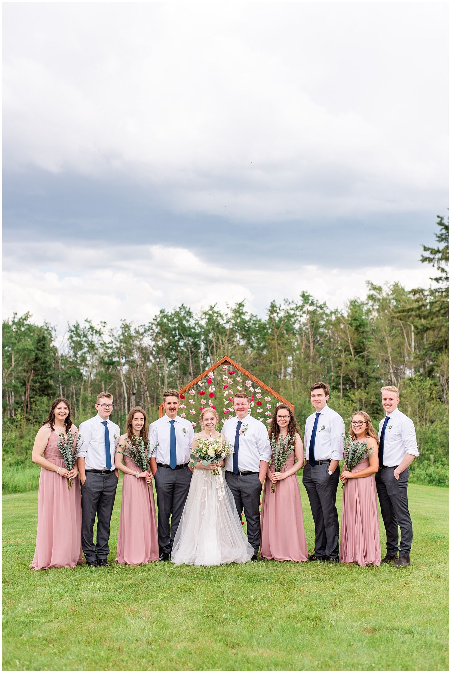 Bridal party of eight stands before a floral arch backdrop on a lush green lawn on a cloudy day.