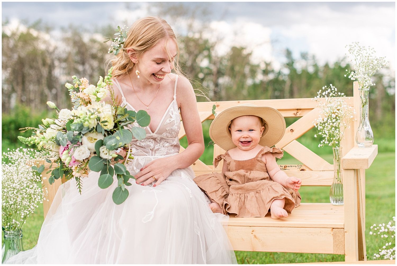 Smiling bride in lace gown sitting on wooden bench with laughing baby in sun hat outdoors.