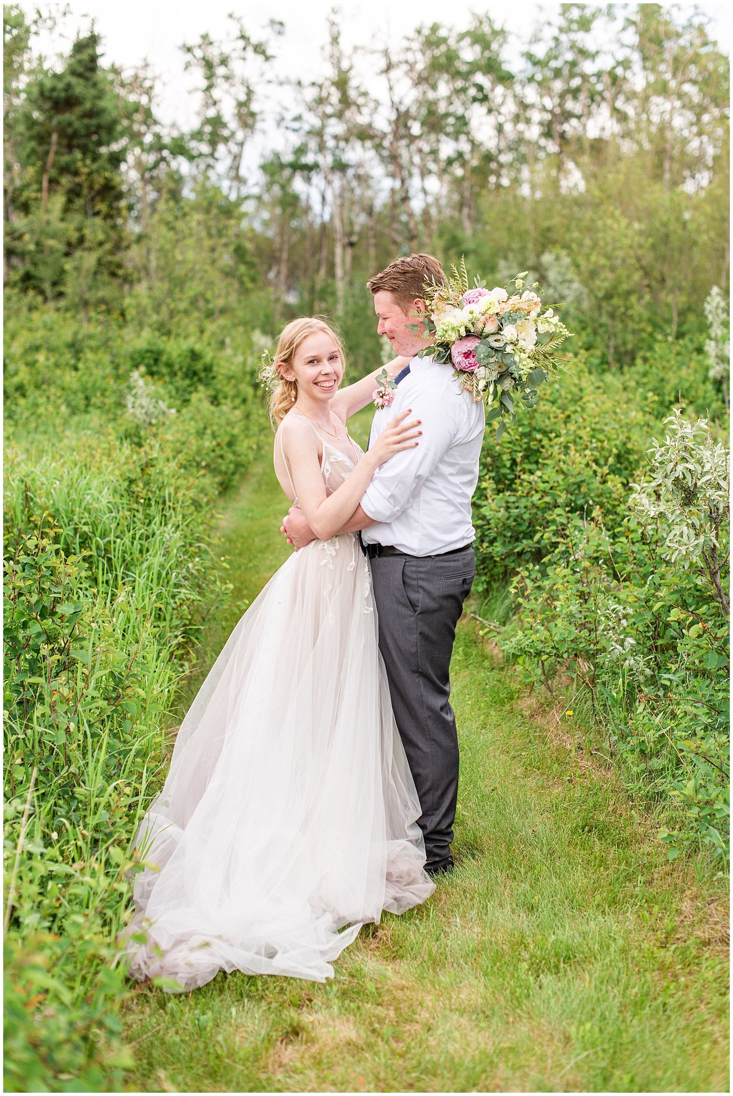 Bride and groom embrace in a lush green garden path, bride wearing a flowing blush tulle gown holding a pastel bouquet.