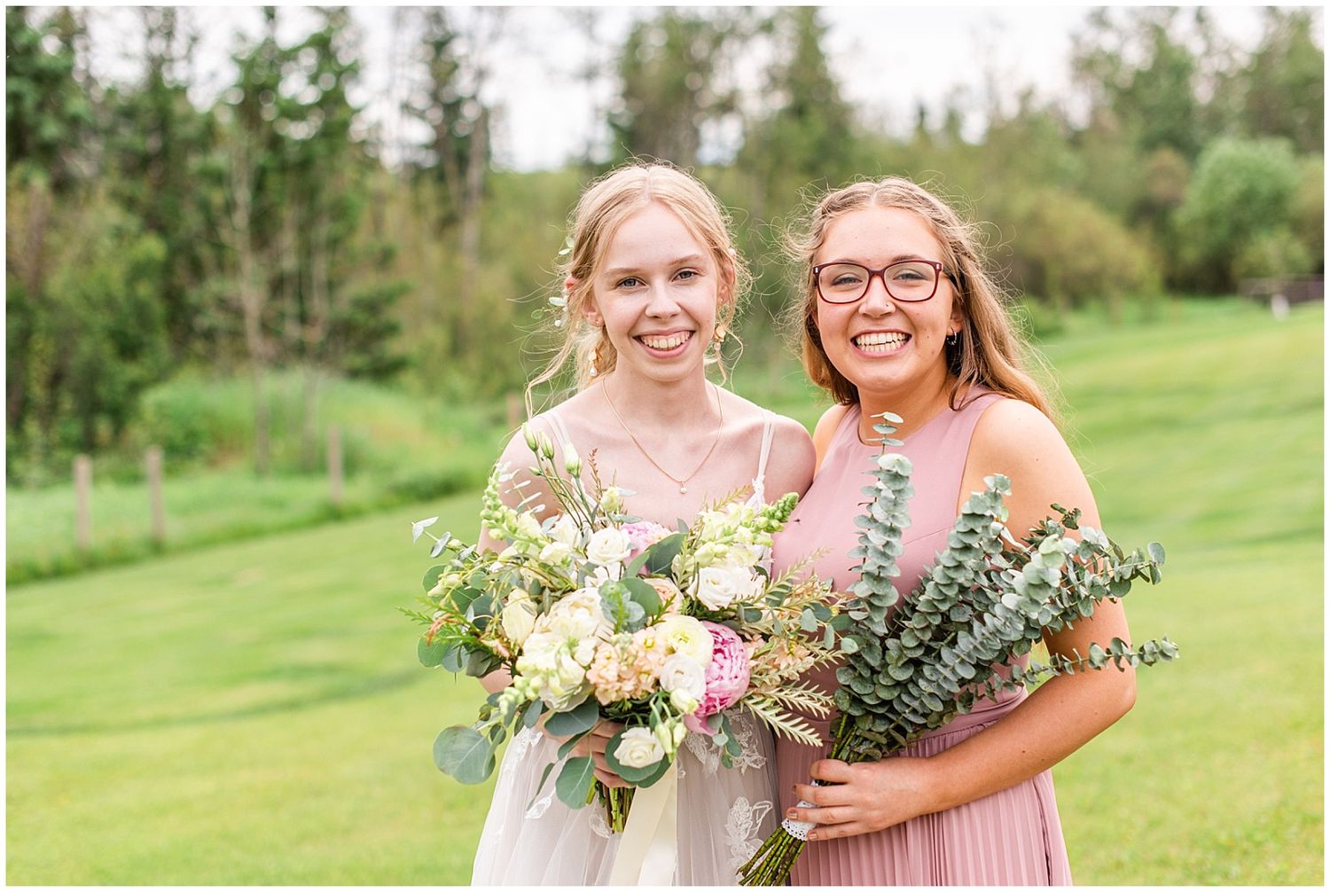 Two bridesmaids in blush dresses smile while holding floral bouquets with greenery outdoors at a wedding.