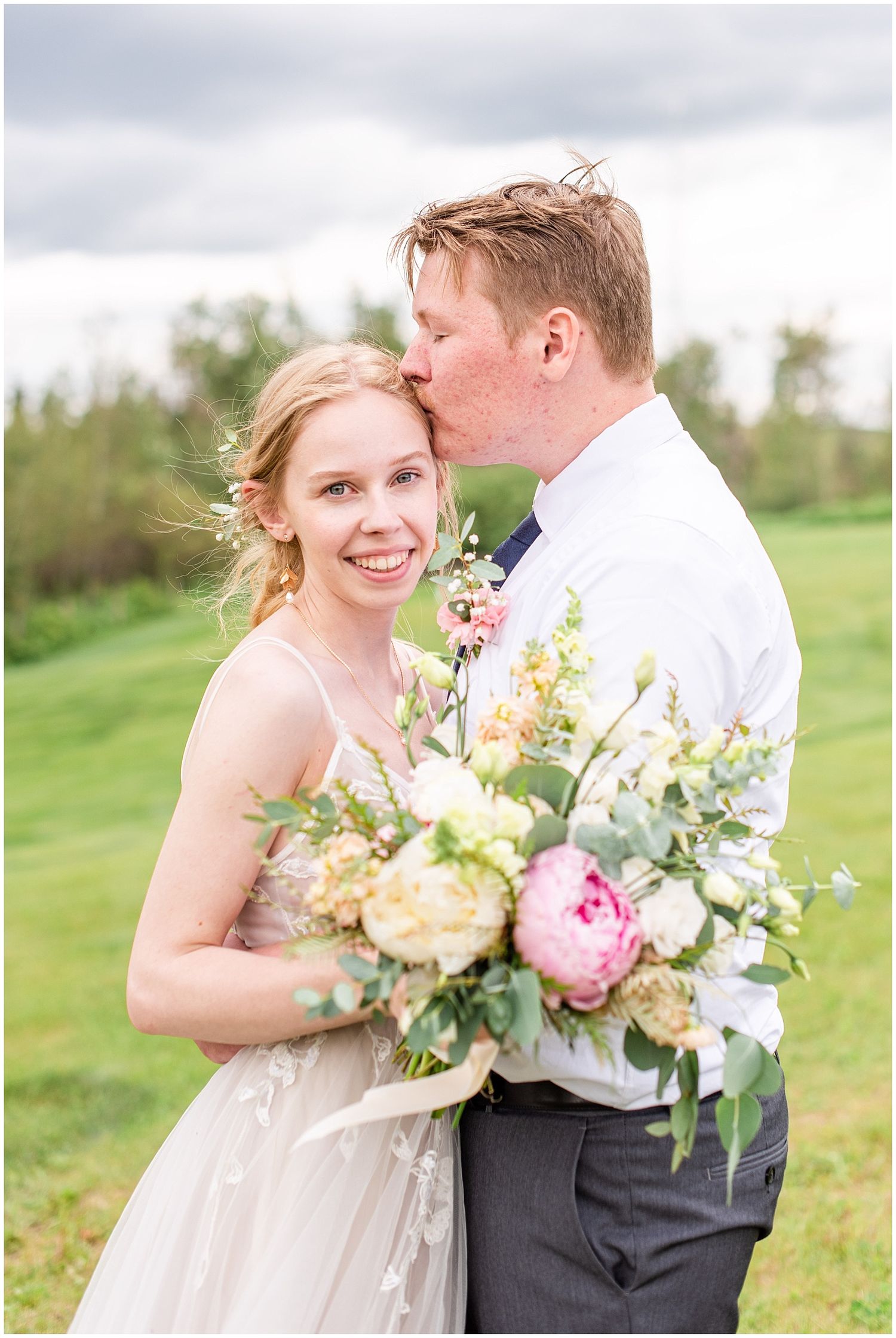 Groom kisses bride on cheek as she smiles holding a lush pink and white peony bouquet outdoors.