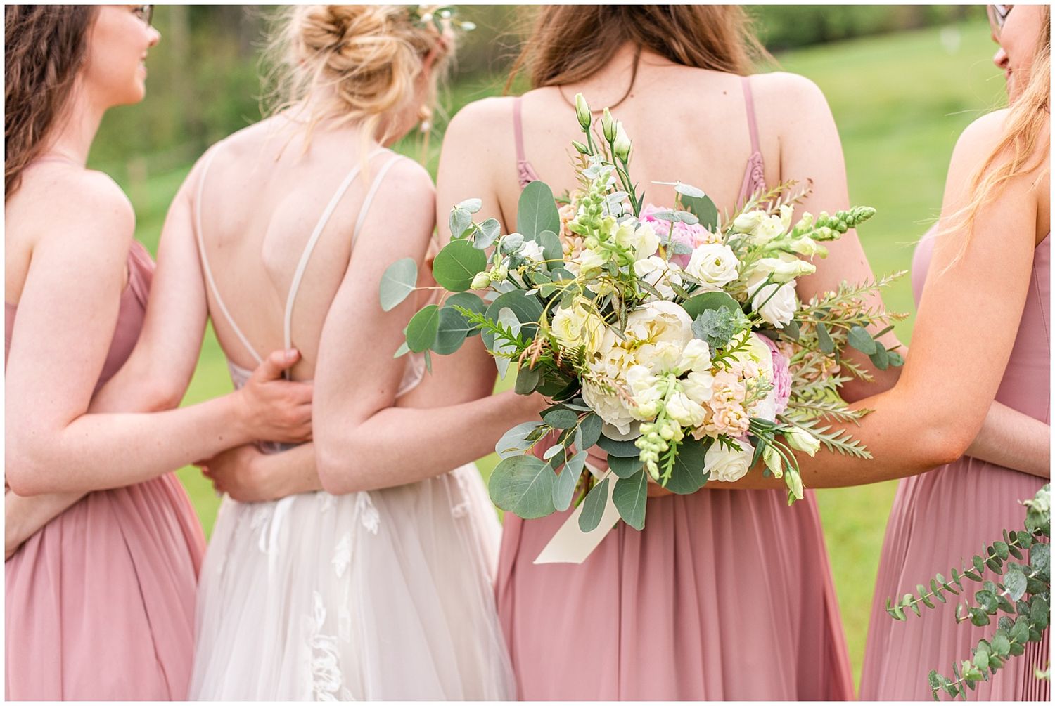 Bridesmaids in dusty rose gowns stand with bride showing elegant open backs and a lush floral bouquet.