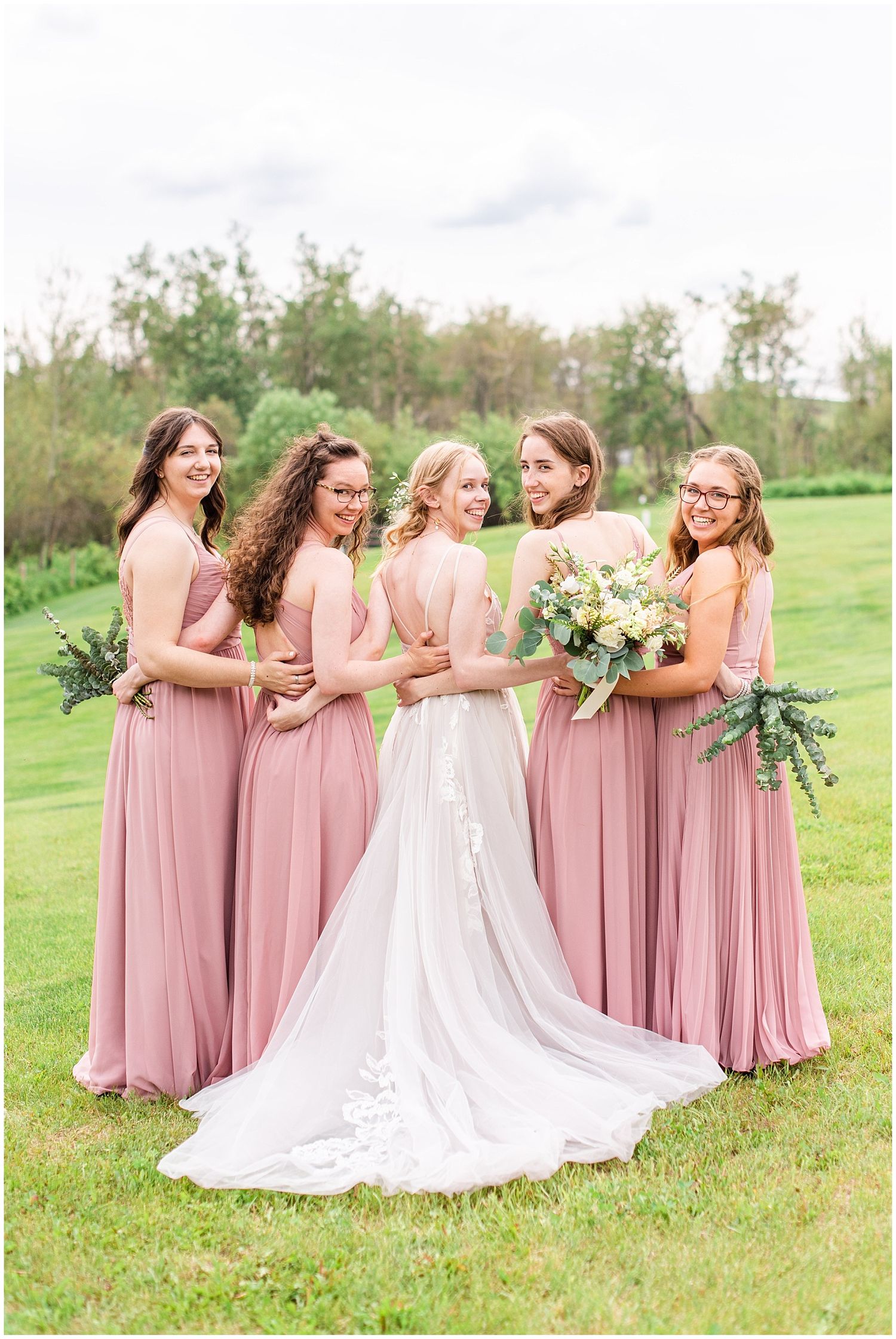 Bride in white lace gown poses with four bridesmaids in dusty rose dresses holding bouquets on a green lawn.