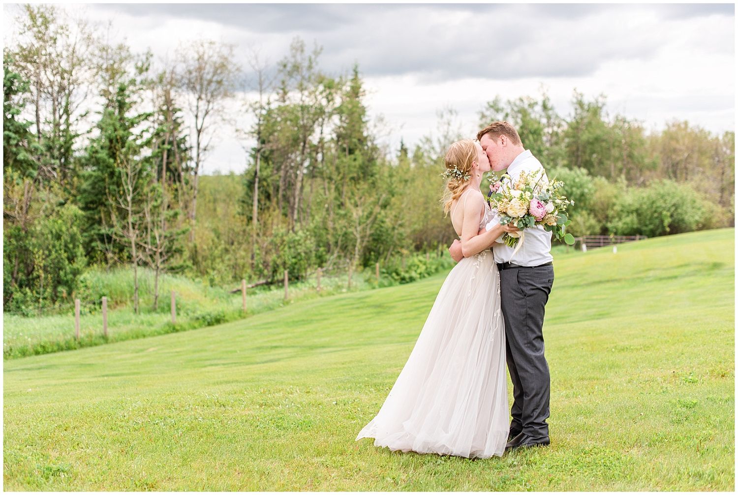 Bride and groom sharing a romantic kiss on a lush green lawn surrounded by trees on their wedding day.