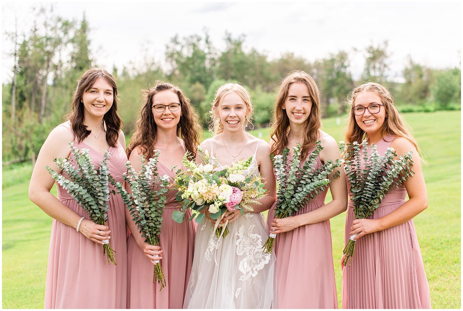 Bride in lace gown poses with four bridesmaids in dusty rose dresses holding bouquets of greenery outdoors.