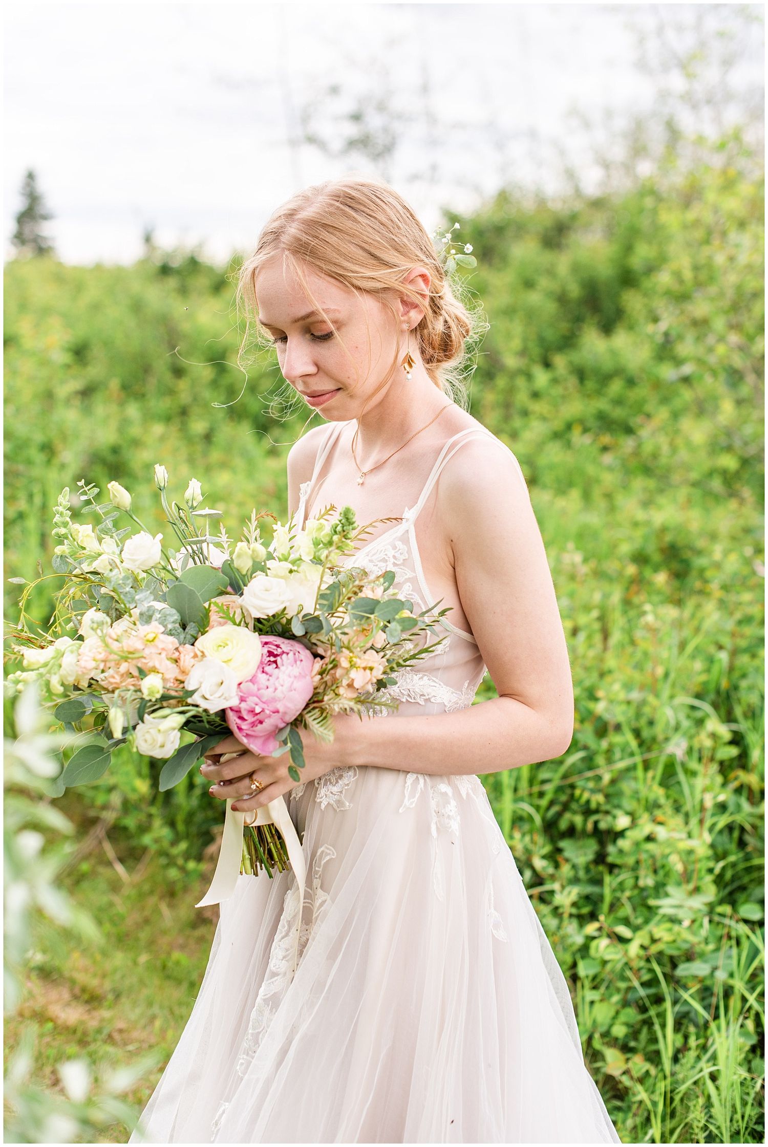 Blonde bride in a delicate lace gown gazes down at her pink and white peony bouquet surrounded by lush greenery.