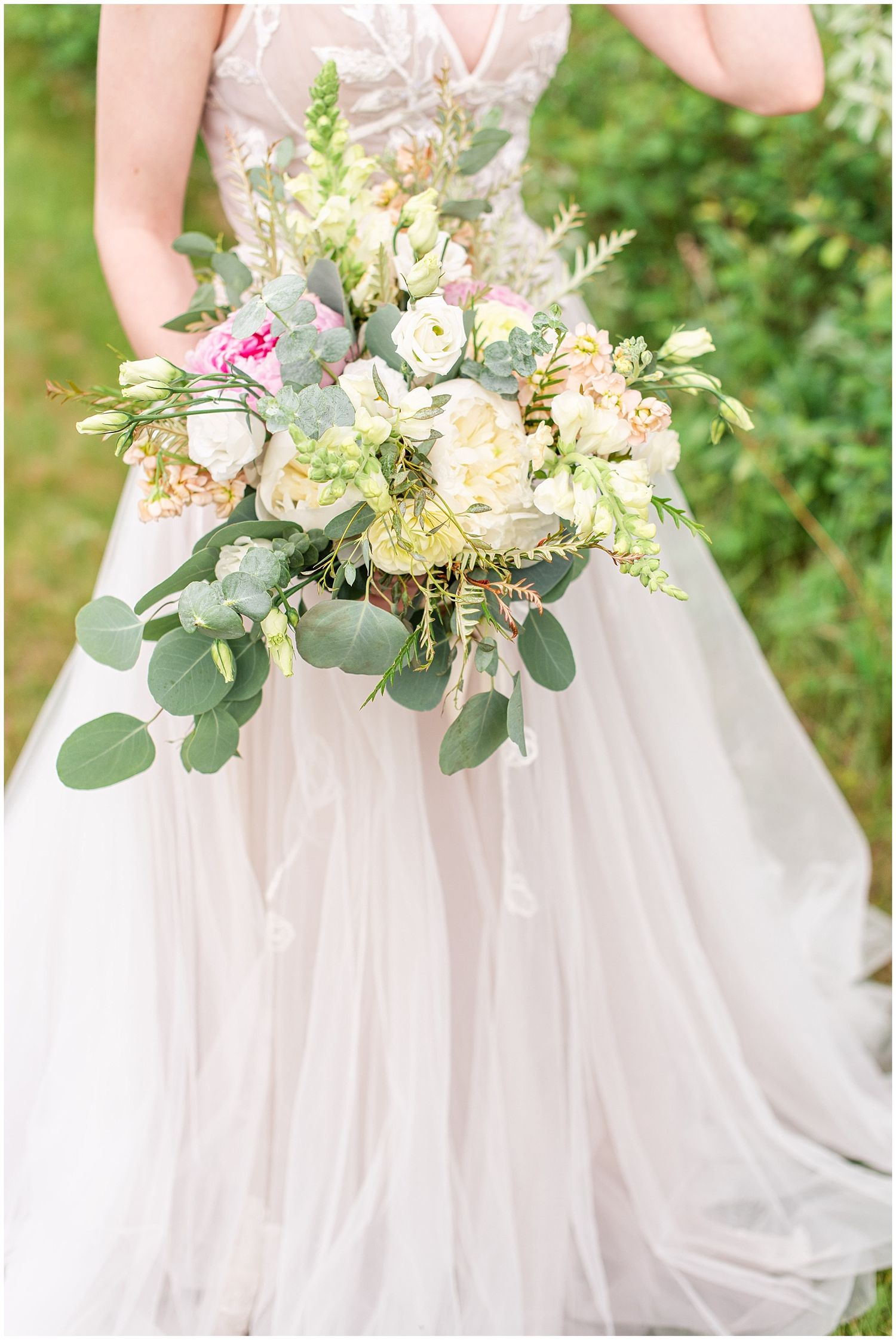 Bride holding a lush wedding bouquet with cream, pink roses and eucalyptus, wearing a soft blush tulle gown outdoors.