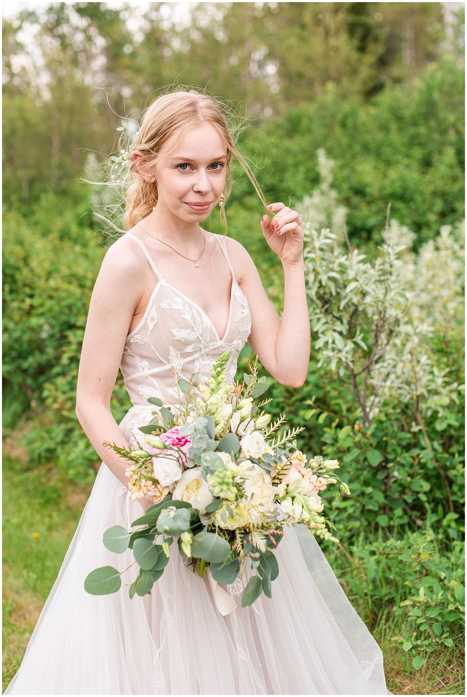 Blonde bride in a lace tulle wedding gown holding a lush bouquet of flowers in a garden setting.