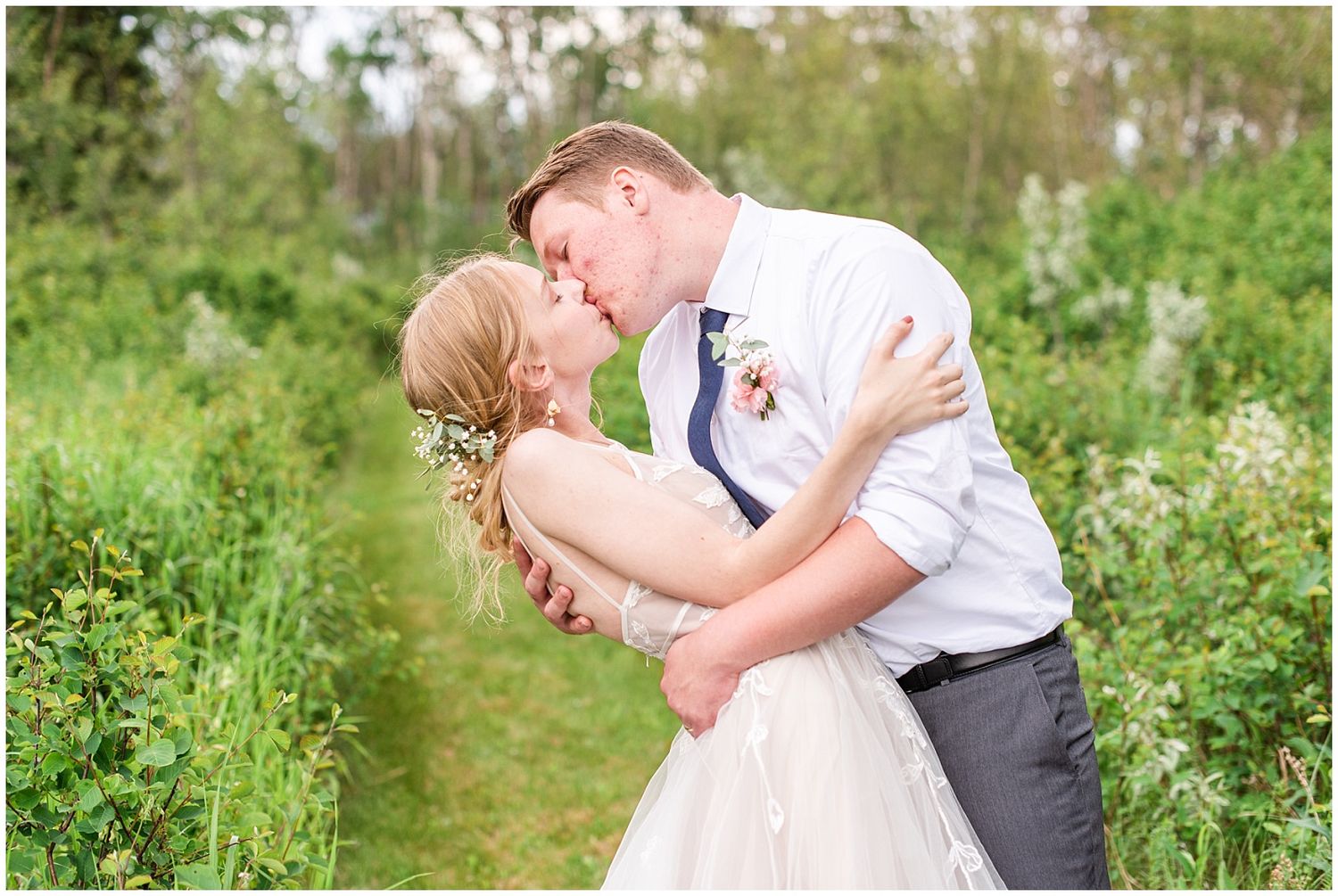 Groom dips bride for a passionate kiss surrounded by lush green foliage in an outdoor garden setting.