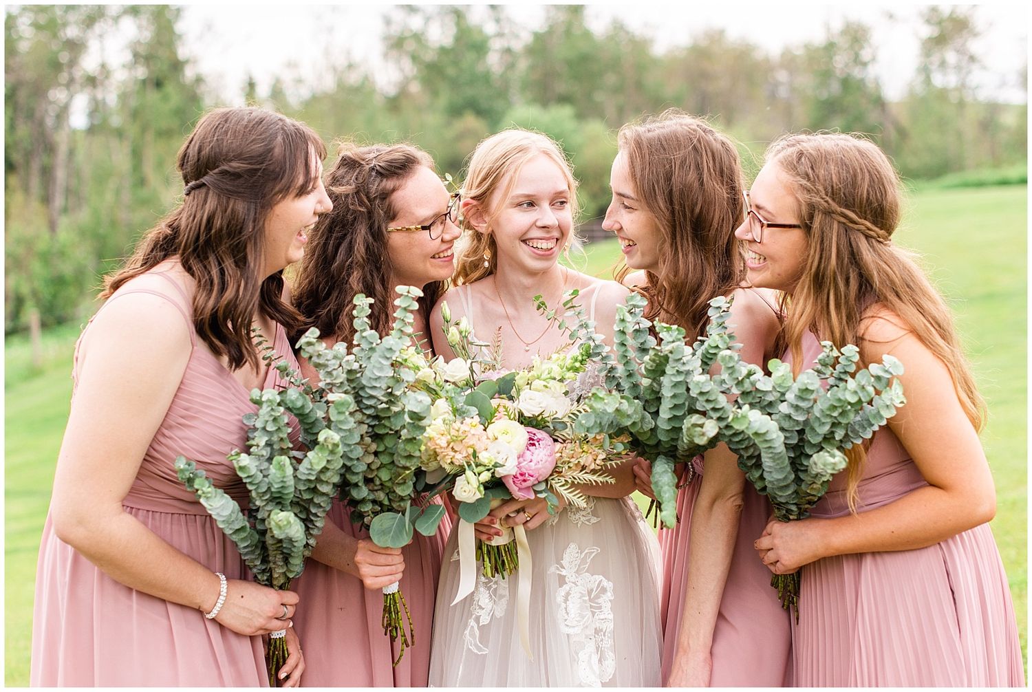 Bride laughs joyfully with four bridesmaids in dusty rose dresses holding eucalyptus and floral bouquets.