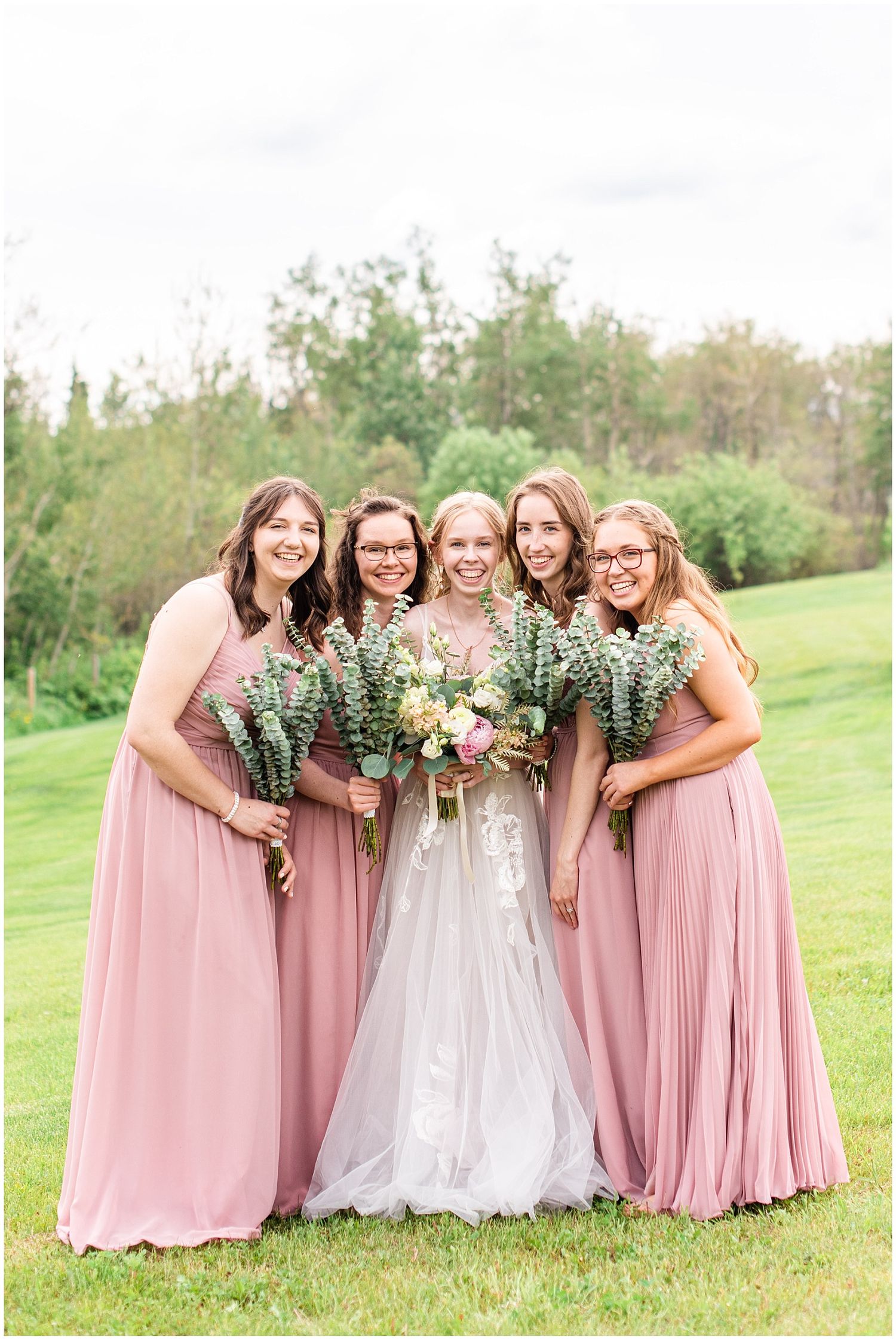 Bride in white gown poses with four bridesmaids in dusty rose dresses holding eucalyptus bouquets outdoors.