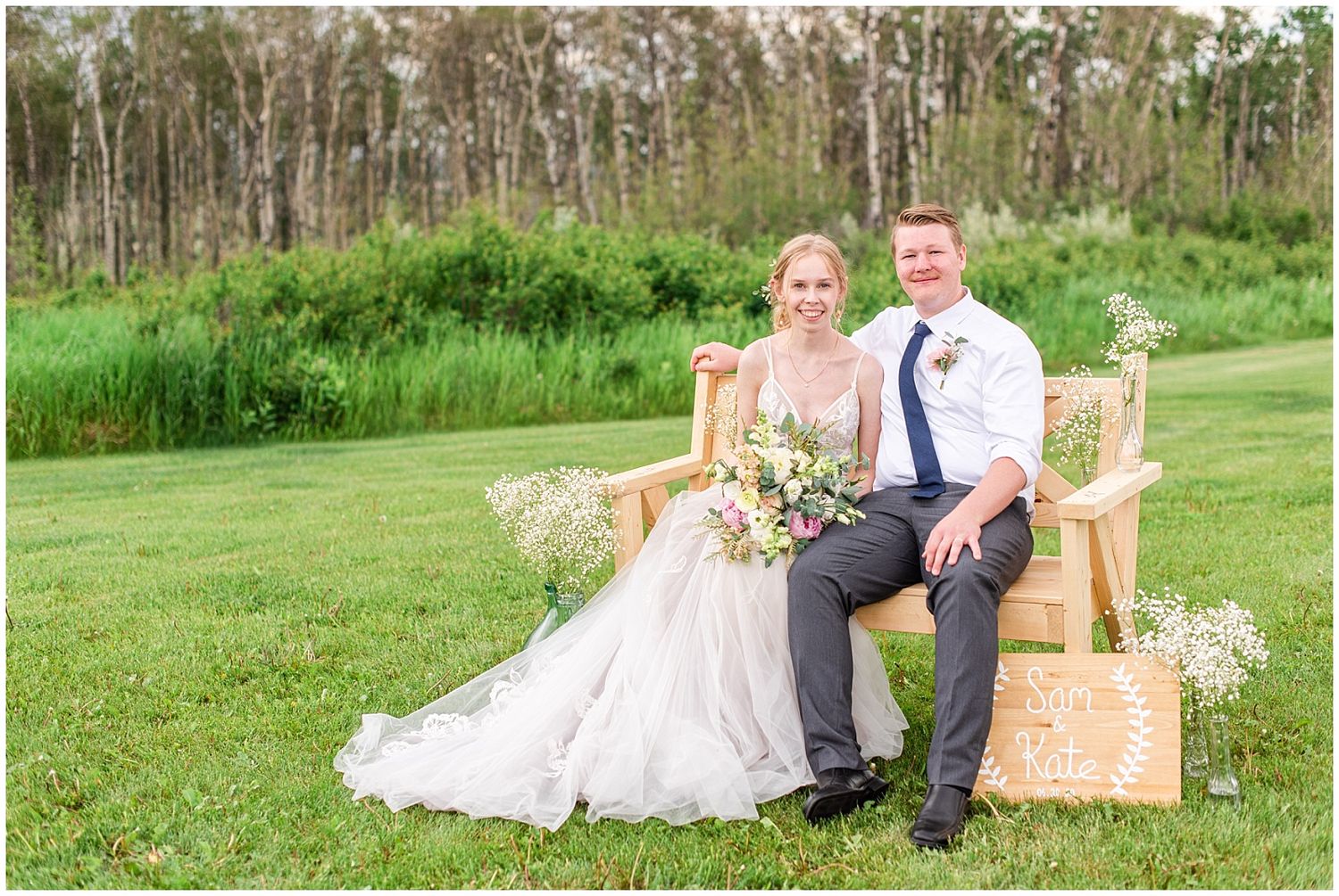 Bride and groom sit together on a wooden bench surrounded by wildflowers and green meadows on their wedding day.