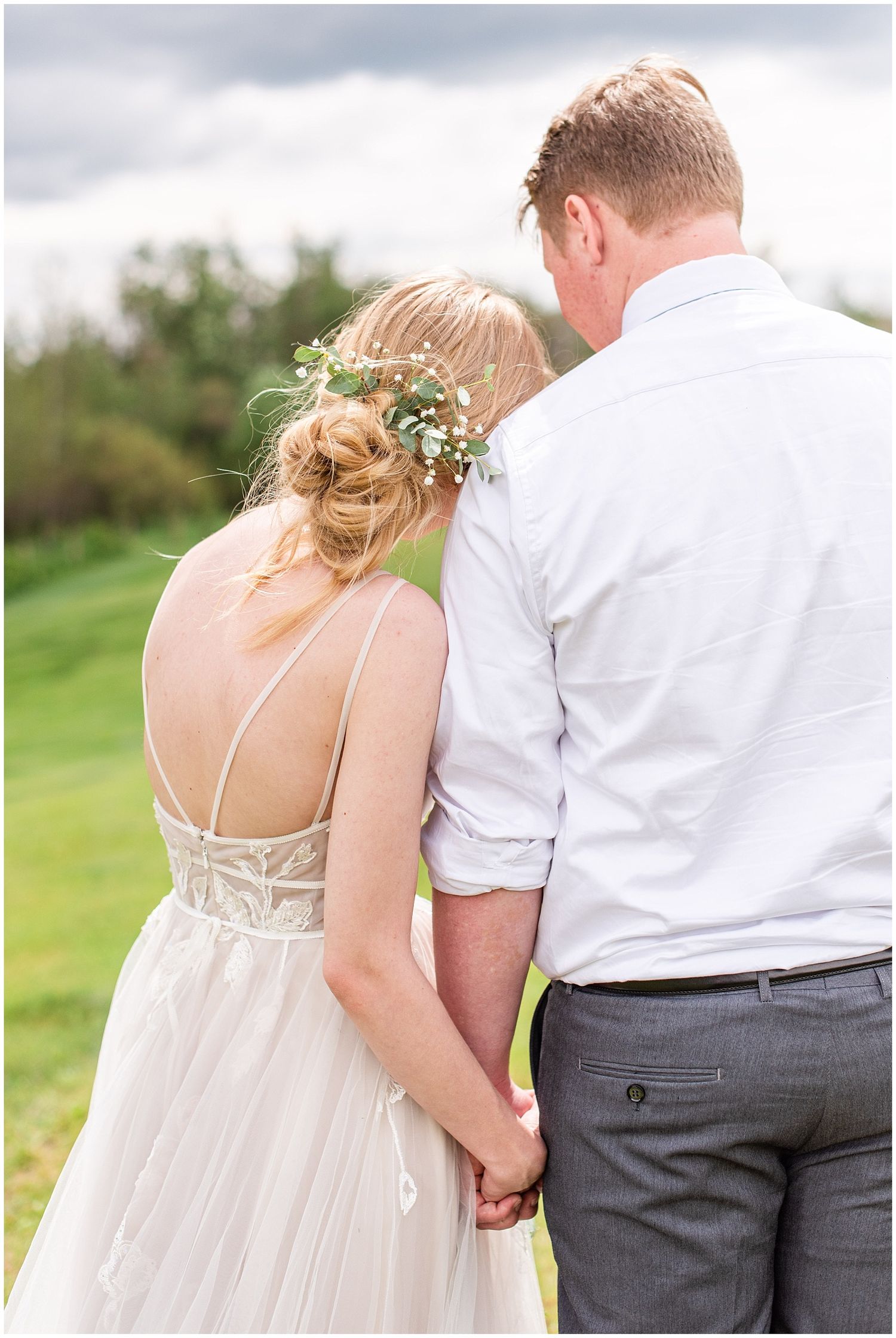 Bride and groom holding hands from behind, she wears a backless gown with floral hair piece, he wears white shirt.