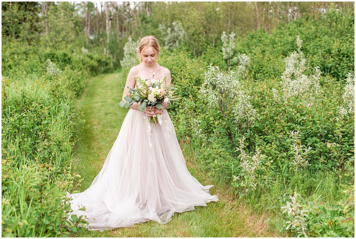 Bride in flowing white gown holds bouquet amid lush green garden path on her wedding day.