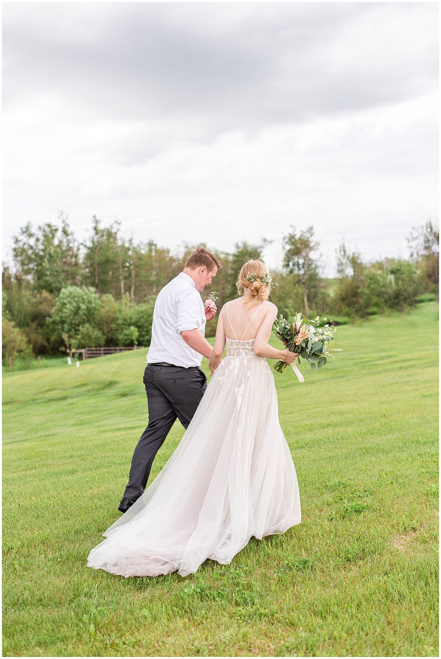 Bride and groom walk hand in hand across a green lawn, bride in a flowing backless gown holding a bouquet.