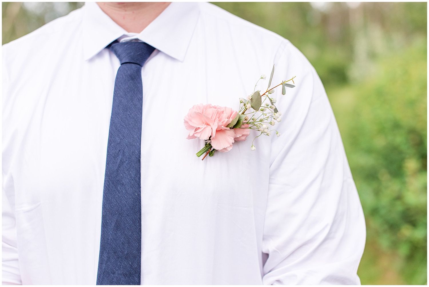 Close-up of a pink floral boutonniere with greenery pinned to a groom's white shirt with a navy tie.