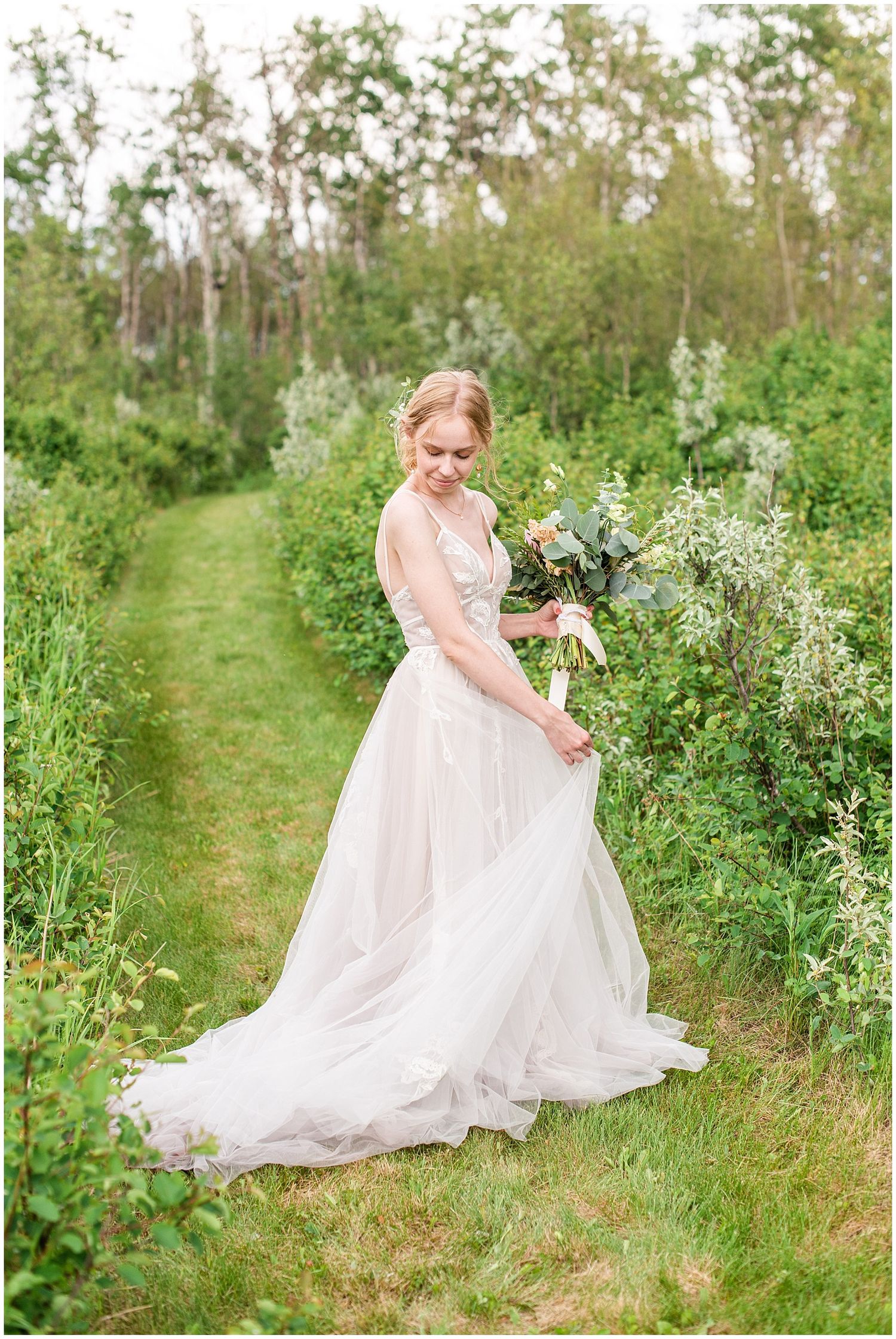 Bride in a flowing tulle gown holding a bouquet on a green garden path surrounded by lush foliage.