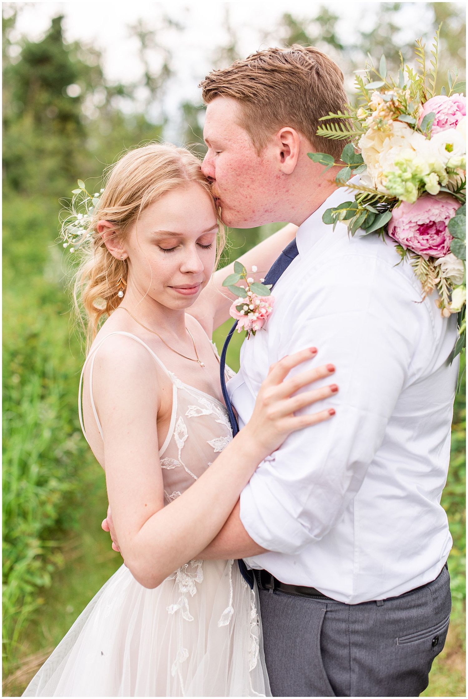 Groom kissing bride on forehead outdoors, bride in floral crown holding bouquet of pink and white flowers.