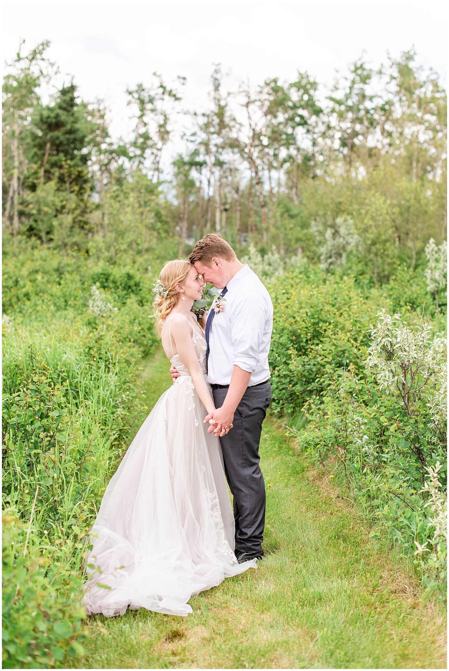 Bride and groom share tender forehead touch while holding hands in scenic green garden pathway on wedding day.