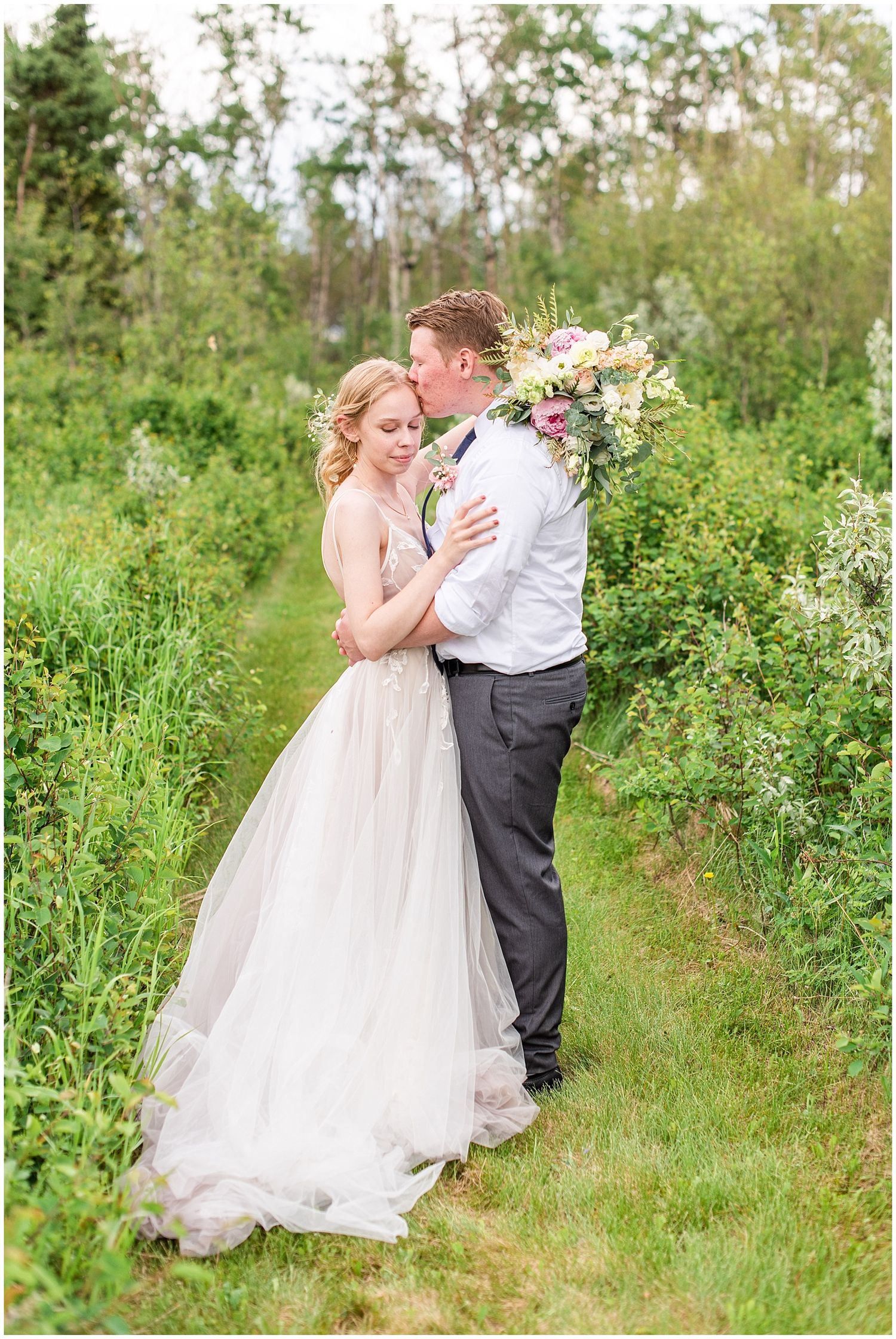 Bride and groom share a tender kiss among lush greenery, bride holding a colorful bouquet in a flowing gown.