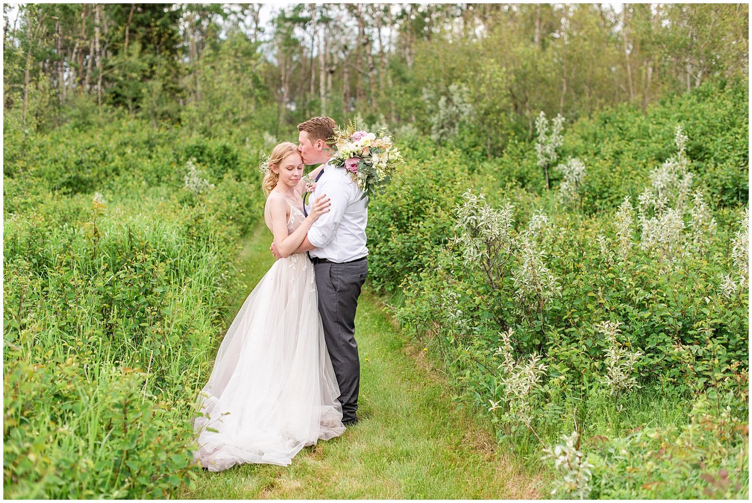 Bride and groom embrace in a lush green meadow, bride holding a wildflower bouquet in a flowing white gown.