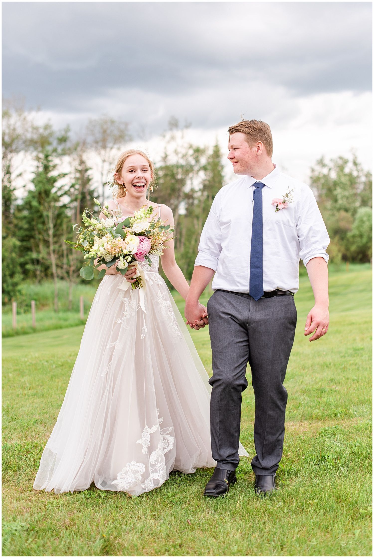 Joyful bride and groom walk hand-in-hand on green grass, bride in lace gown holding a pink floral bouquet.