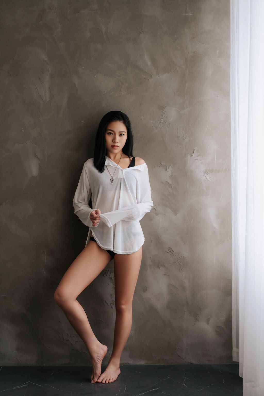 Young Asian woman in white oversized shirt leaning against gray wall, barefoot in minimalist indoor setting.