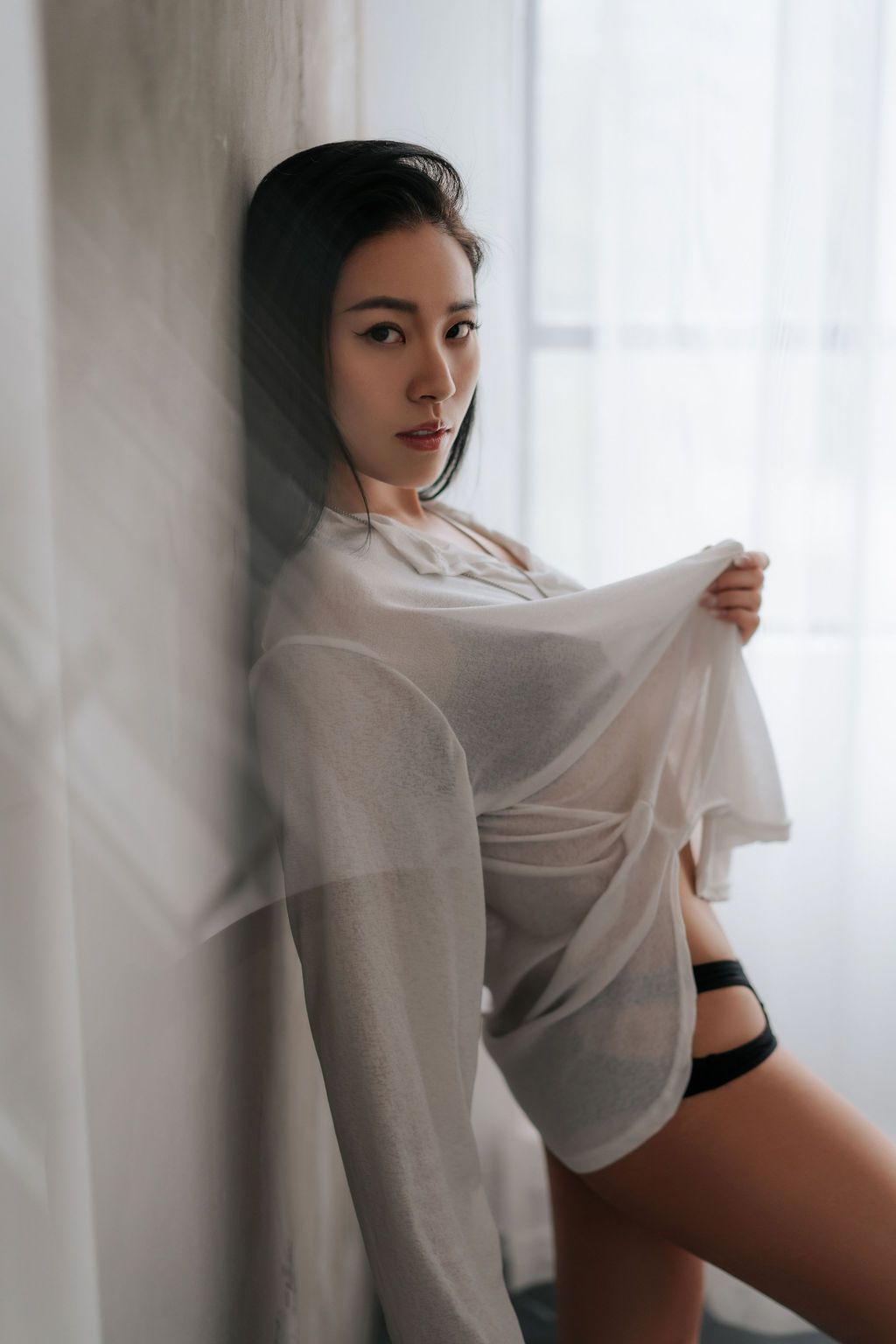 Young woman in sheer white top and black underwear leaning against a wall by a window in soft light.