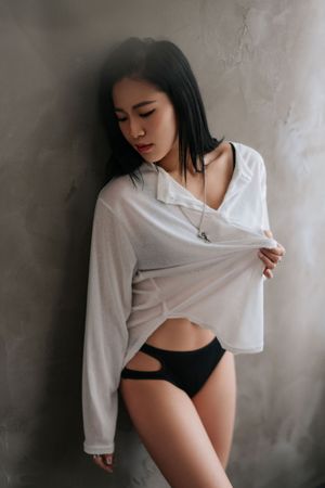 Asian woman in white sheer shirt and black underwear posing against textured gray wall indoors.
