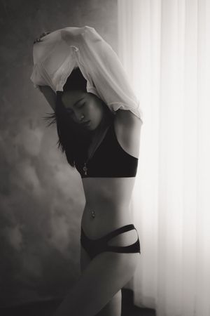 Black and white photo of a woman in lingerie removing a white shirt near a curtained window.