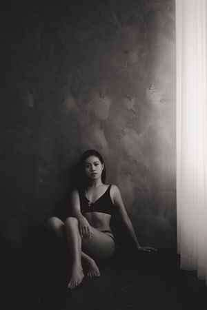 Black and white photo of a young woman sitting on the floor in lingerie against a textured wall near a window.