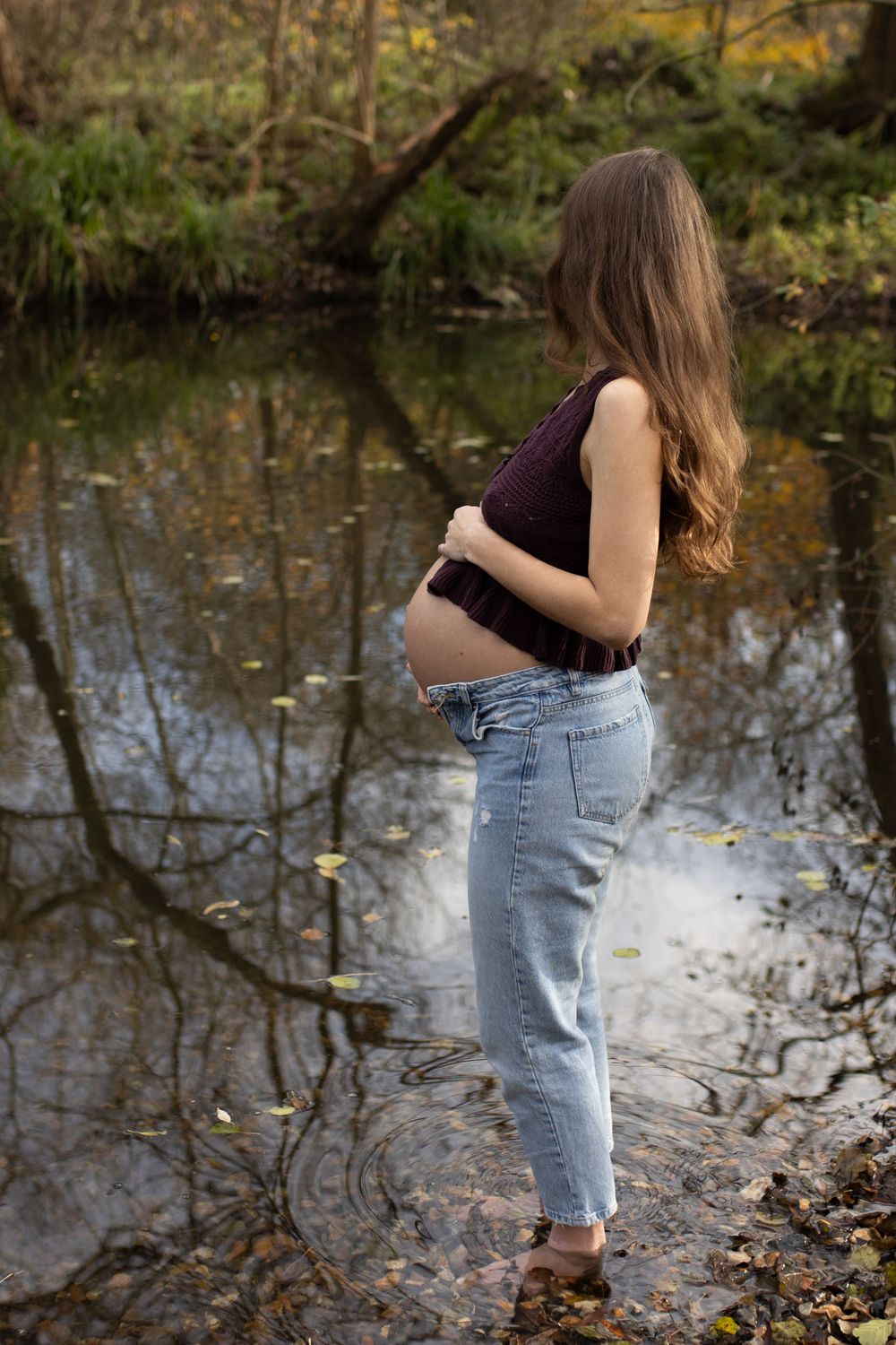 Pregnant woman in jeans and dark top cradling her baby bump beside a reflective forest pond.