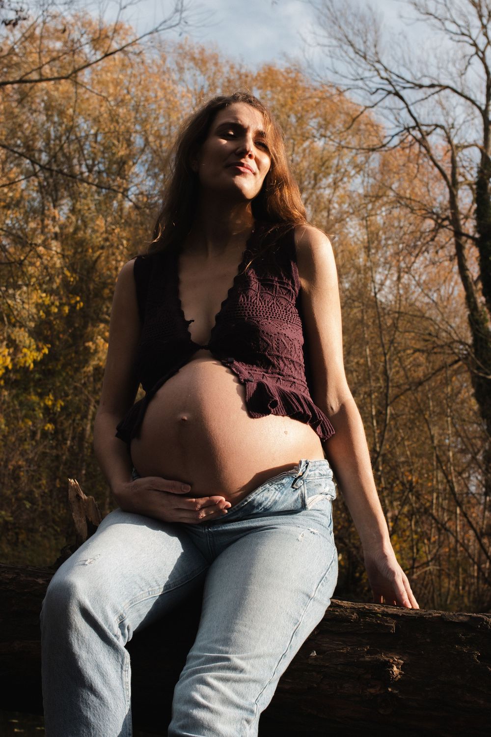 Pregnant woman in jeans and dark top sits on log in autumn forest, cradling her baby bump in golden sunlight.