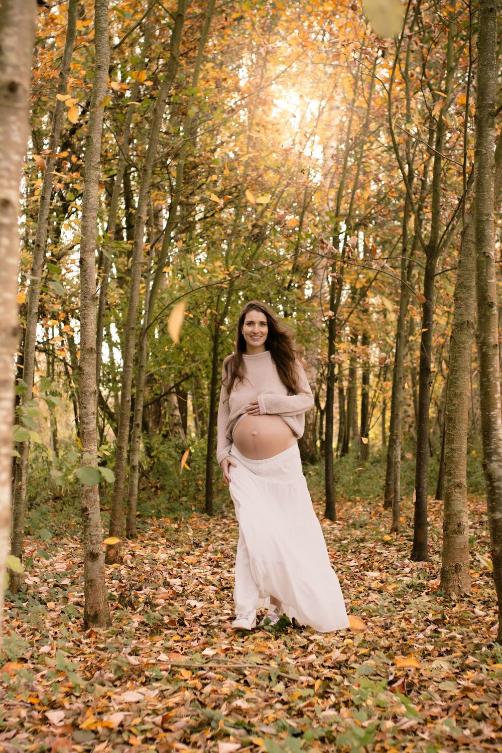 Pregnant woman in beige dress cradling baby bump in autumn forest with golden leaves and warm sunlight.