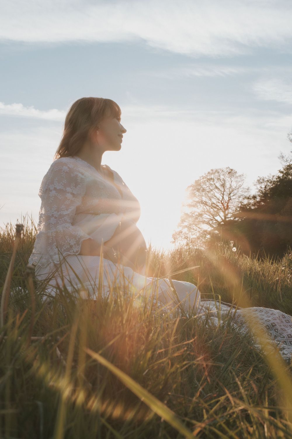 Pregnant woman sitting in golden hour sunlight in a grassy field, wearing a white dress, with sun flare and blue sky.