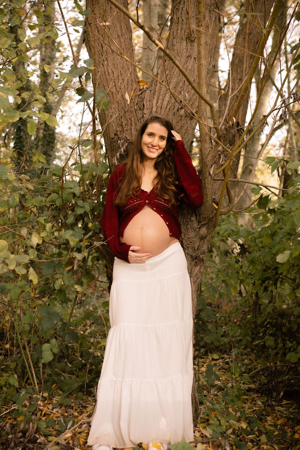 Smiling pregnant woman in white maxi skirt and red cardigan leaning against large tree in lush green forest.