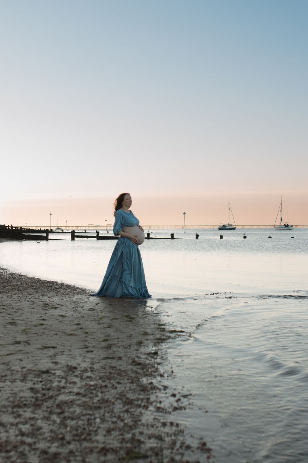 Pregnant woman in blue dress standing in shallow ocean water at sunset with sailboats in background.
