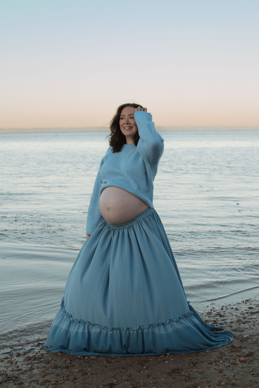 Pregnant woman in blue crop sweater and maxi skirt posing on beach at sunset with ocean backdrop.