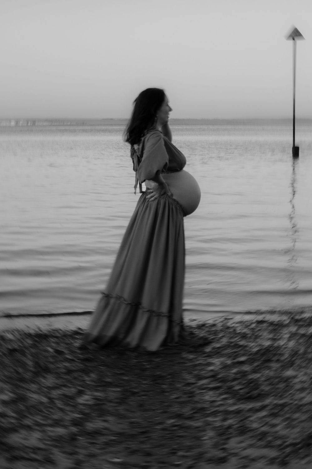 Black and white photo of a pregnant woman in a flowing gown standing by the water at dusk, cradling her belly.