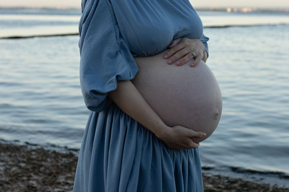 Pregnant woman in blue dress cradling her bare baby bump by the ocean shore at dusk.