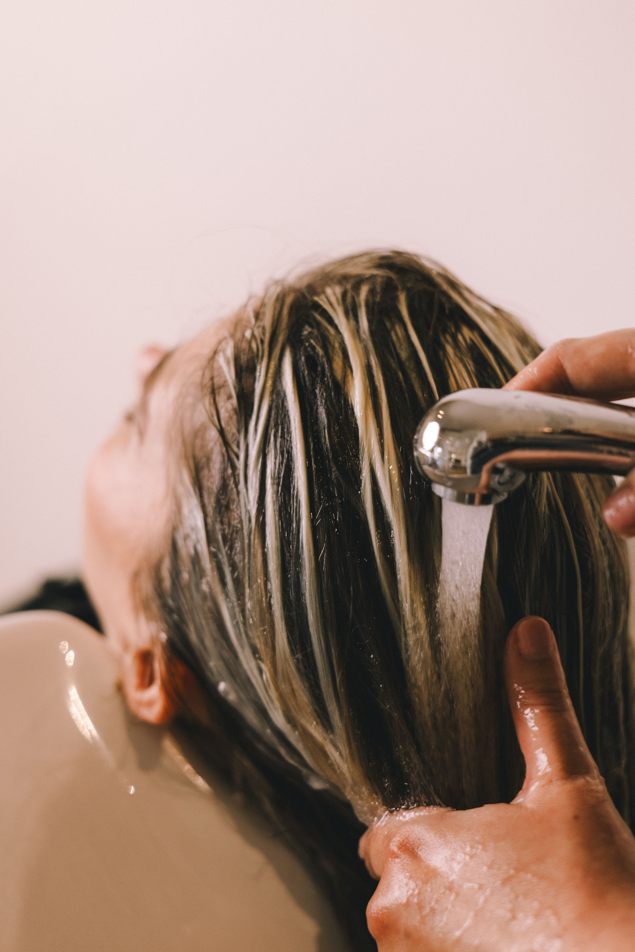 Hairstylist rinsing highlighted blonde hair at a salon wash basin with a handheld showerhead.