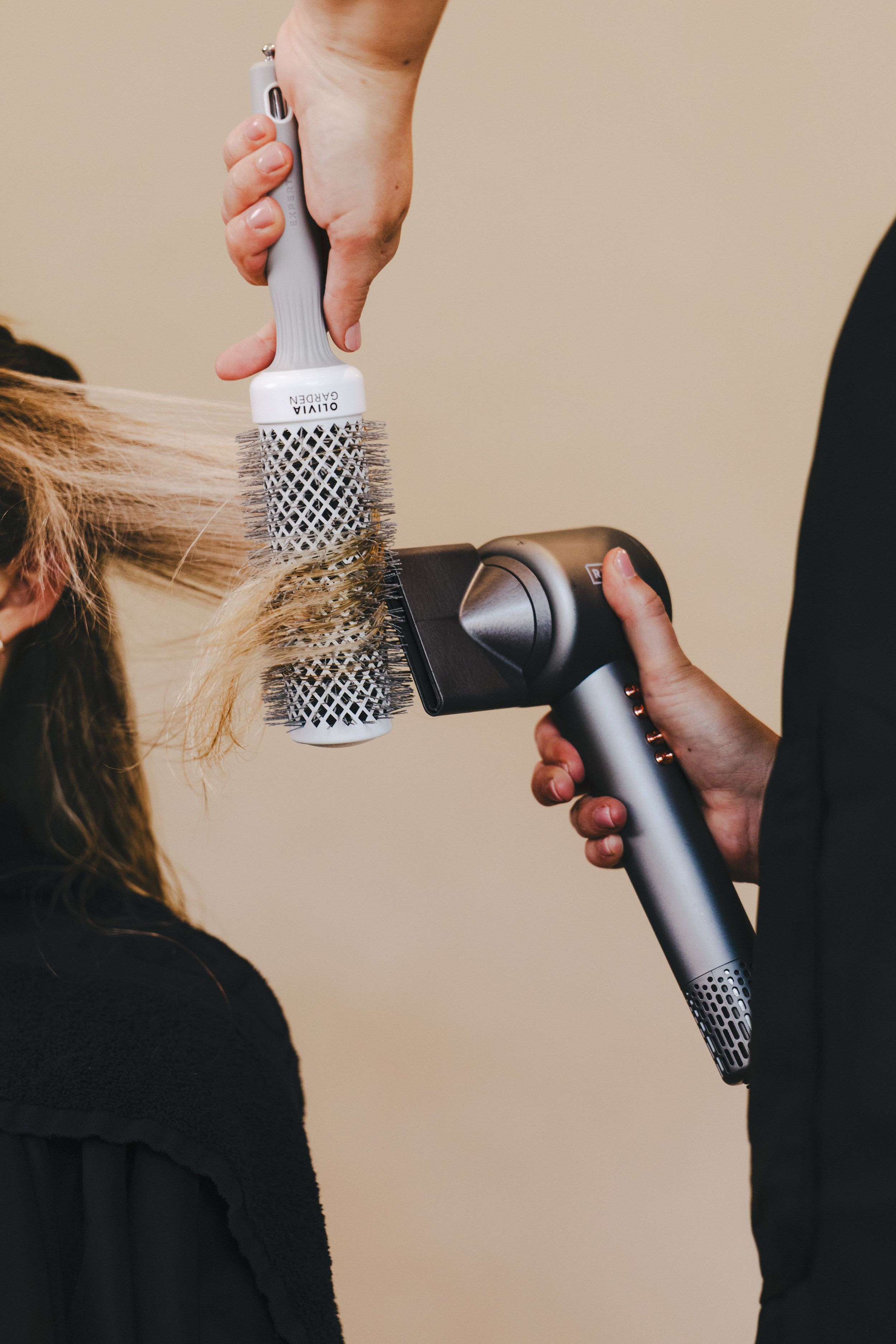 Hairstylist using a round brush and hair dryer to blow dry a client's blonde hair in a salon.