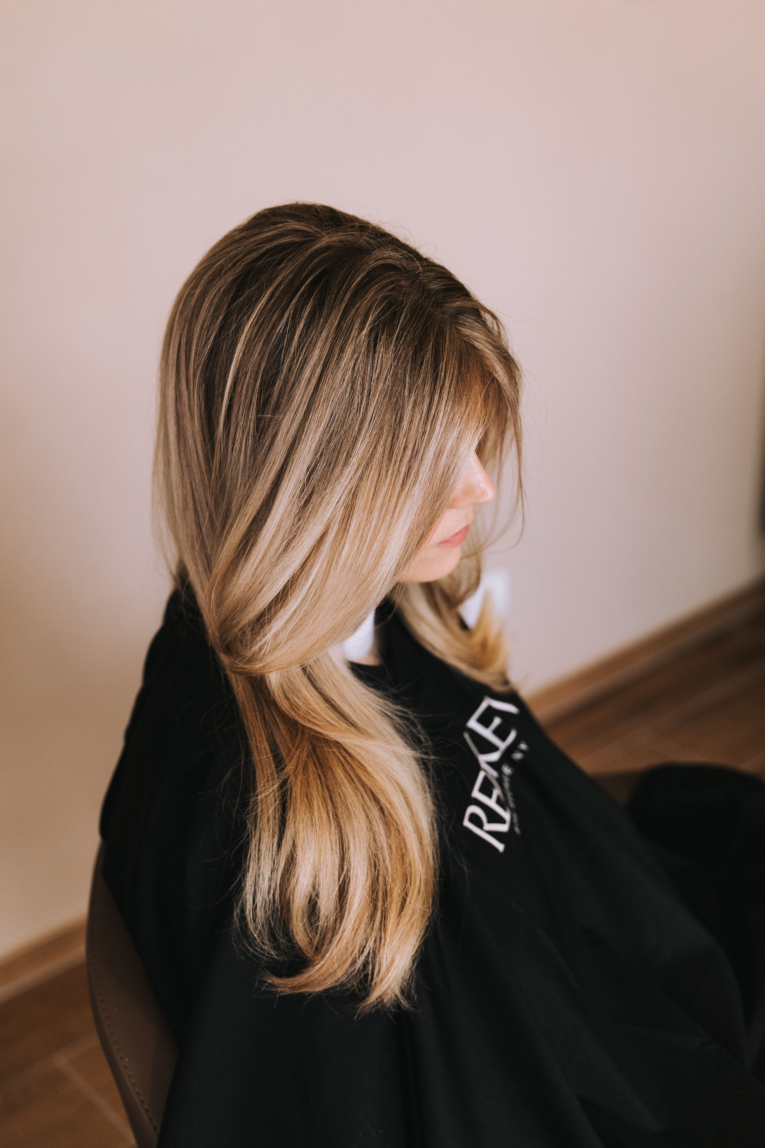 Woman with long blonde balayage hair sitting in salon chair wearing black cape, showcasing highlights.
