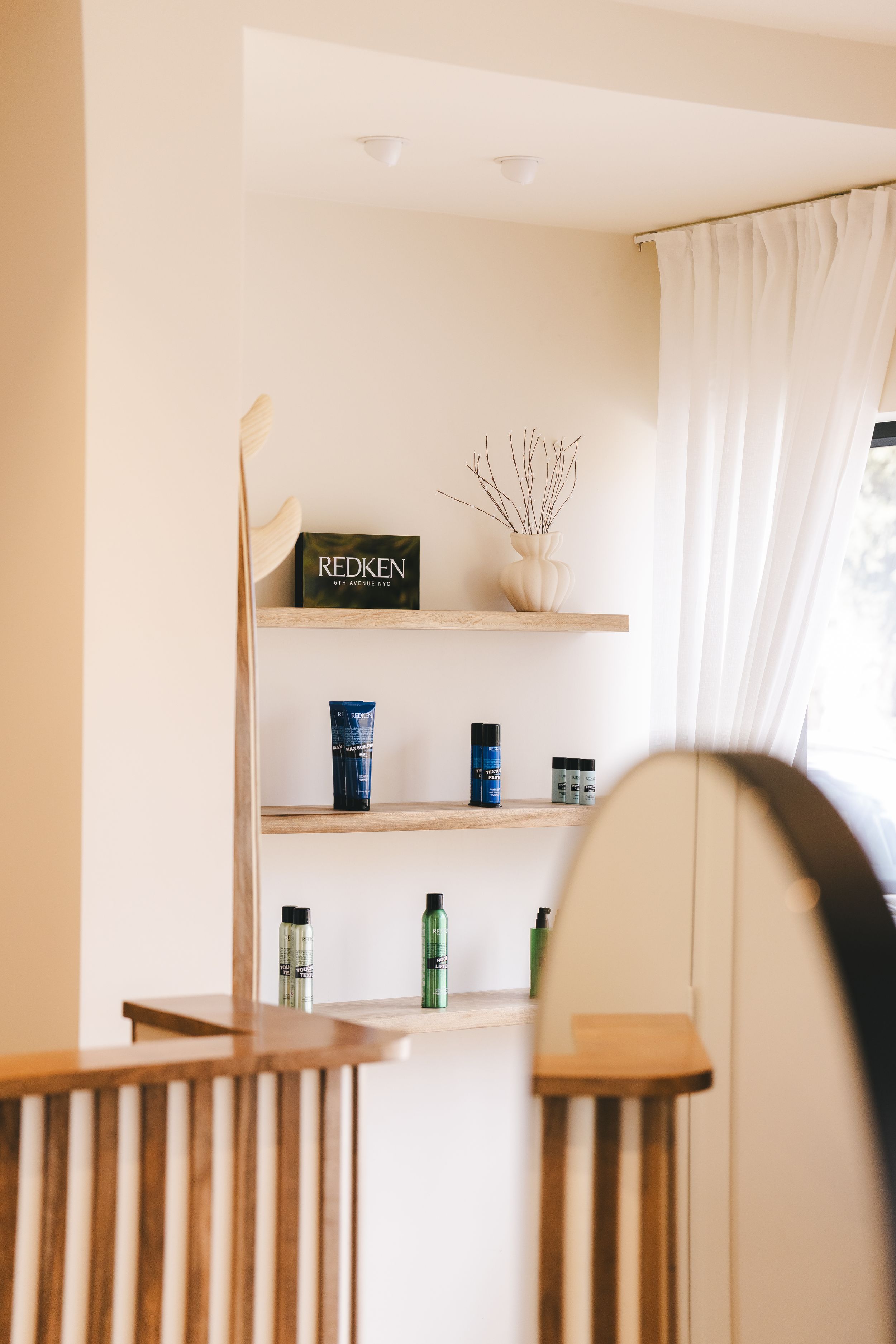 Wooden shelves displaying Redken hair products and a vase near a bright window in a modern salon.