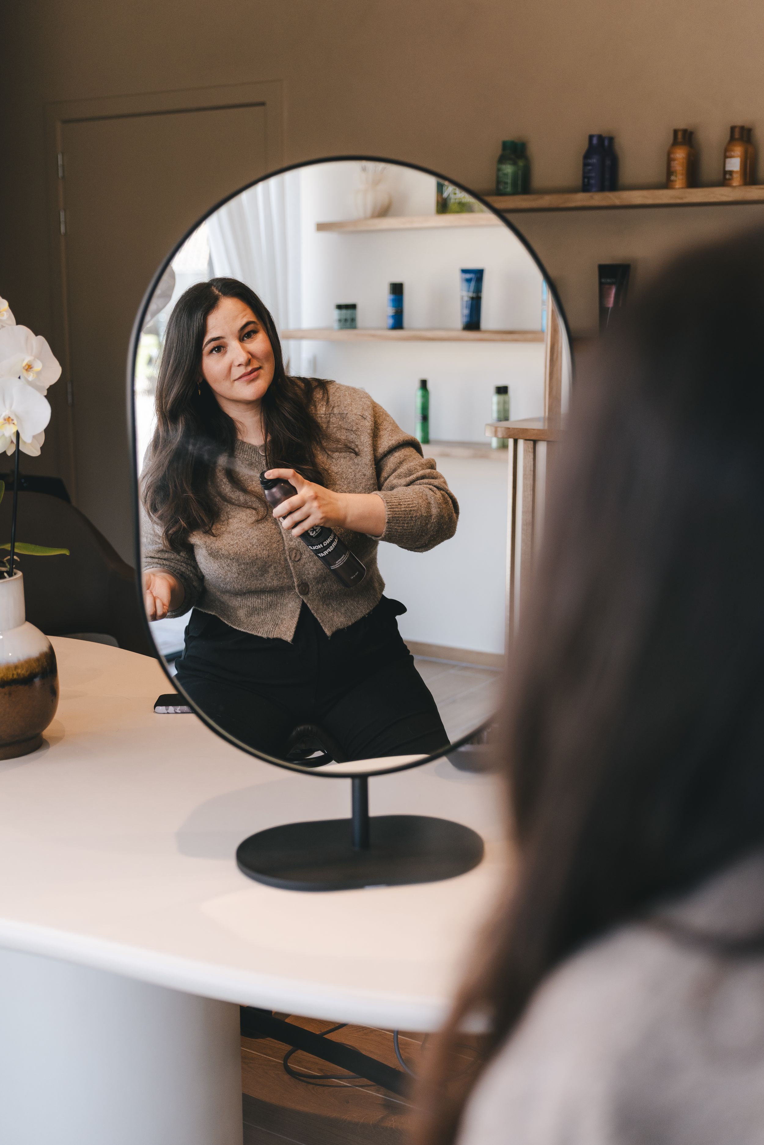 A smiling woman holding a spray bottle reflected in an oval mirror at a modern hair salon.