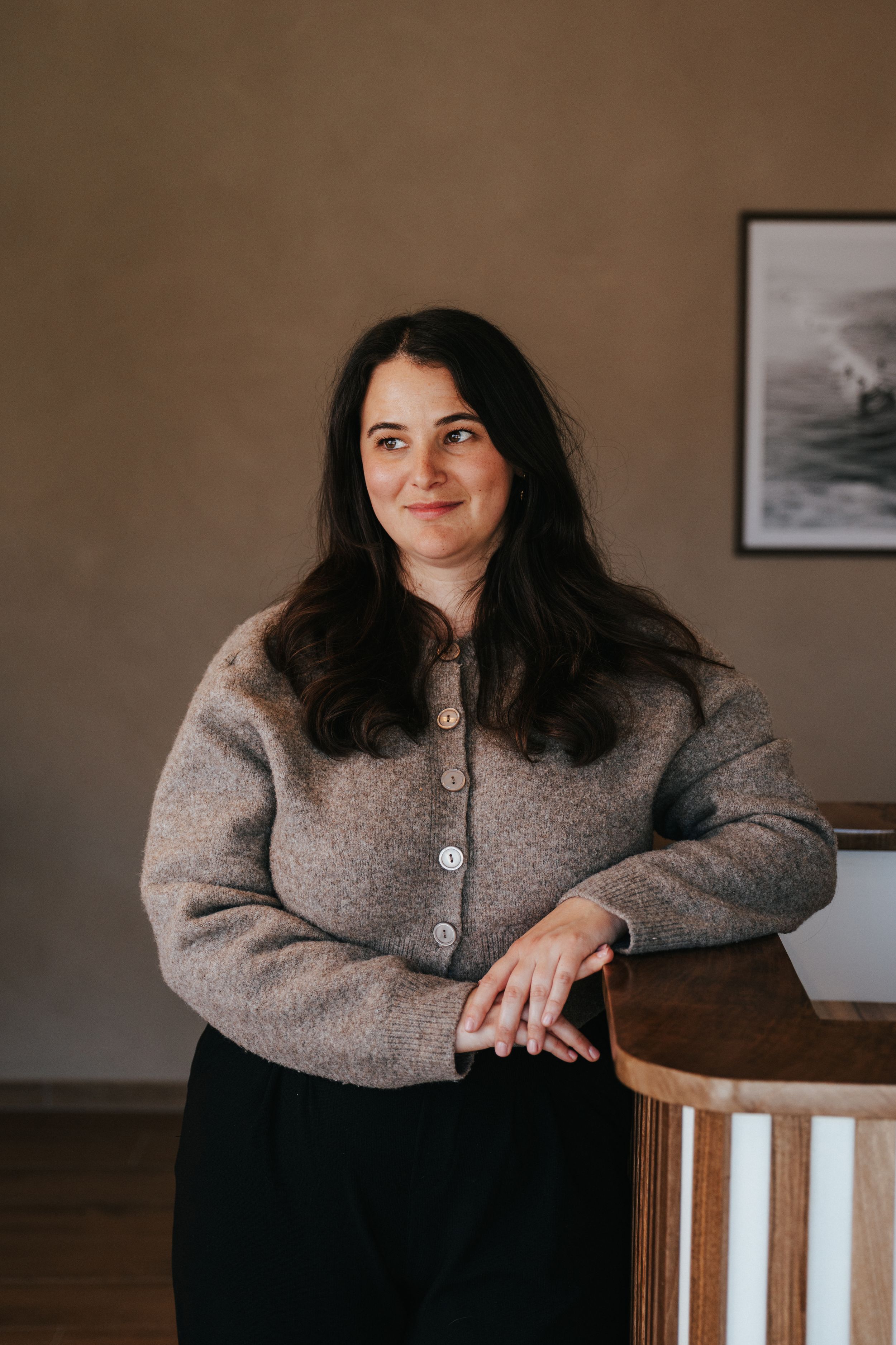 Woman in gray wool cardigan leaning on wooden furniture, smiling in a modern interior space.