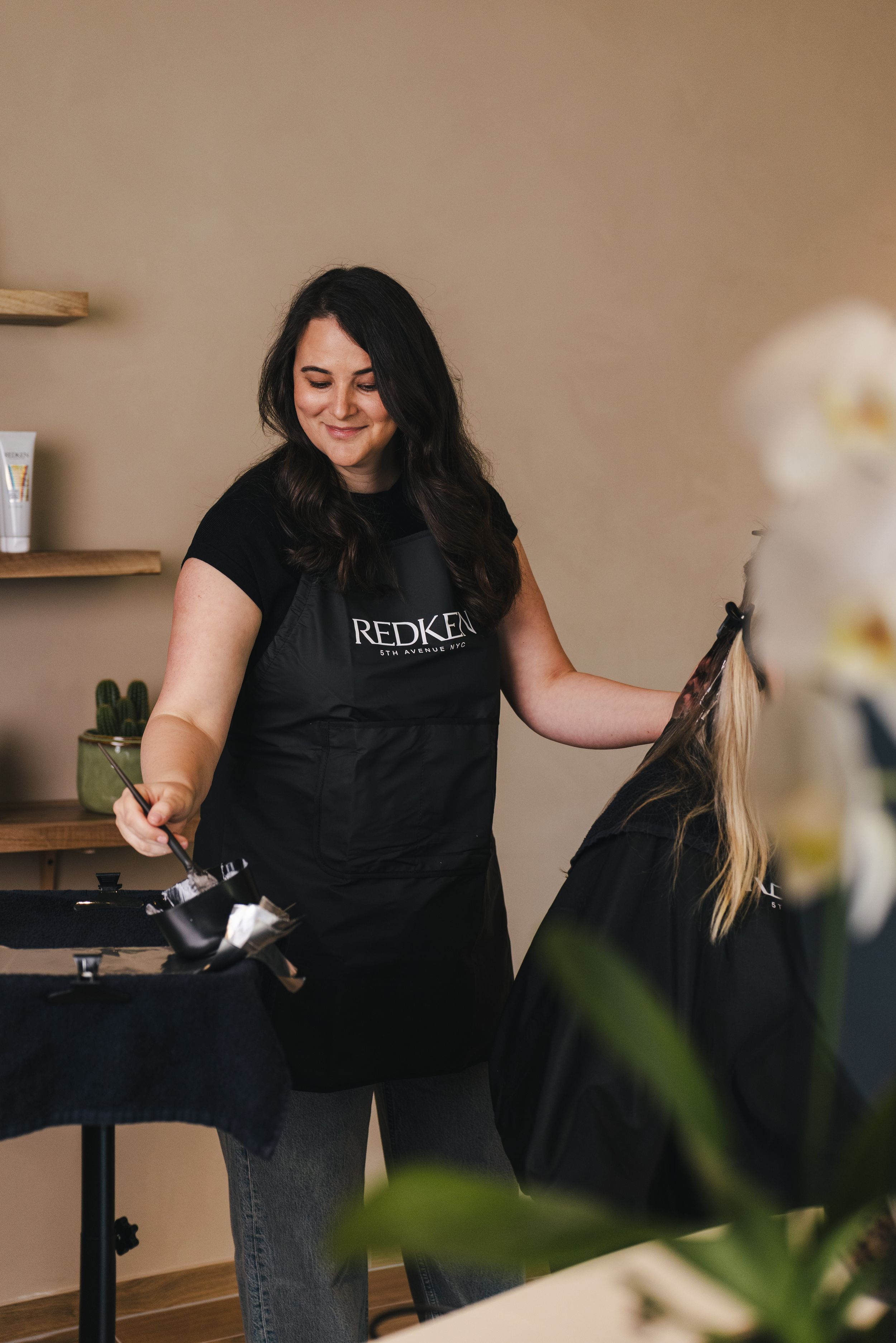 A smiling hairstylist wearing a Redken apron prepares hair color tools at a salon station.