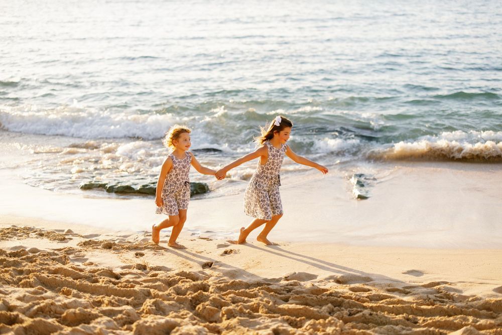 Two young girls in floral dresses hold hands running along a sunny sandy beach with ocean waves behind them.